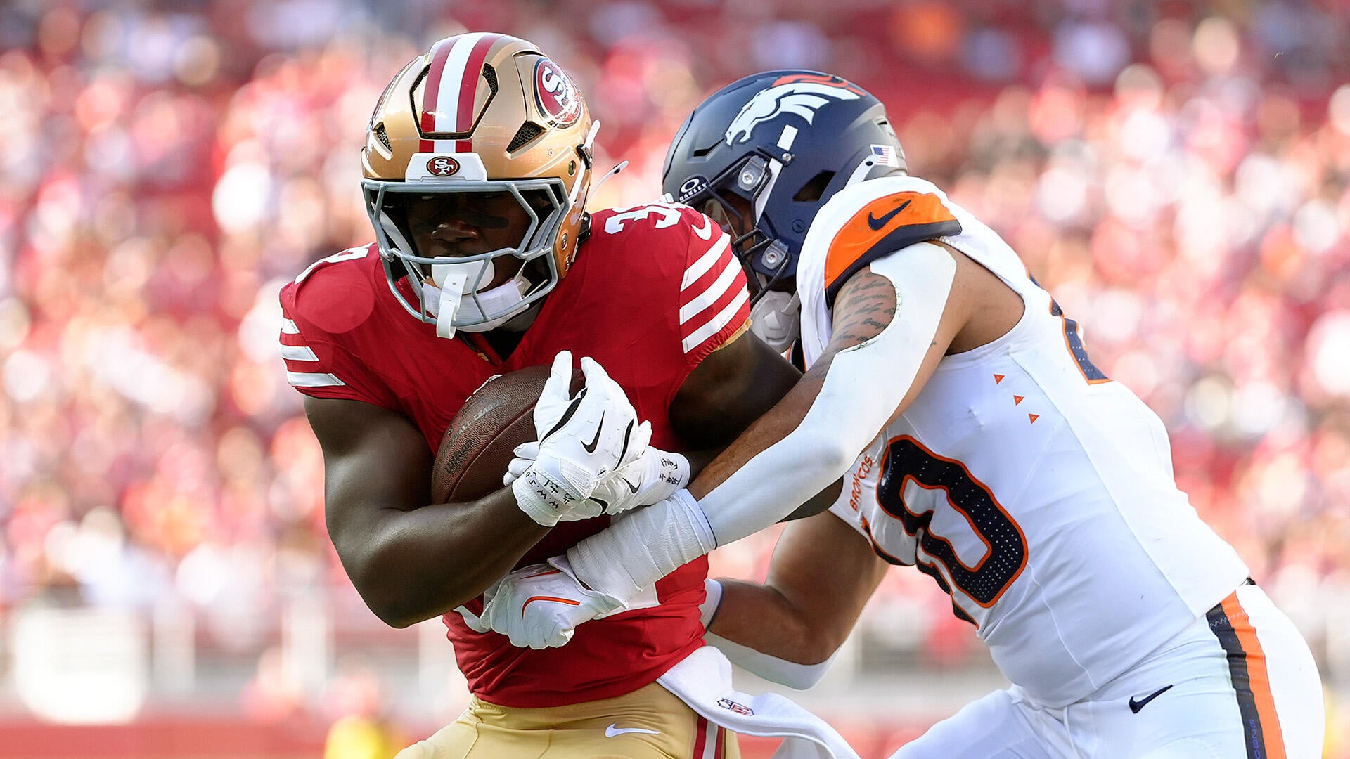 49ers RB, in a red jersey, carries the ball while a Broncos linebacker attempts to tackle him.