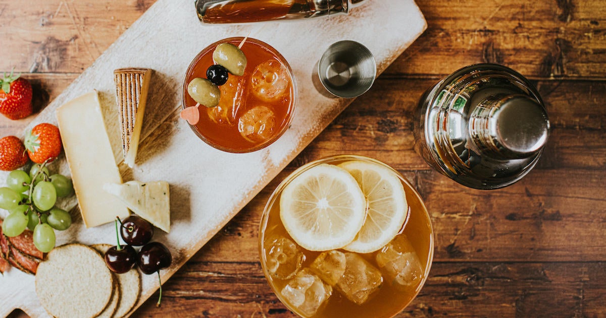 Top-down image of a delicious, simple cheese board and a cocktails on a wooden table beside cocktail shakers