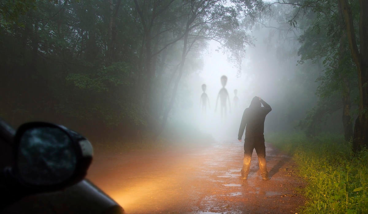 A science fiction concept of a man looking at aliens coming out the mist on a foggy, spooky forest road in the evening. Highlighted by car headlights.