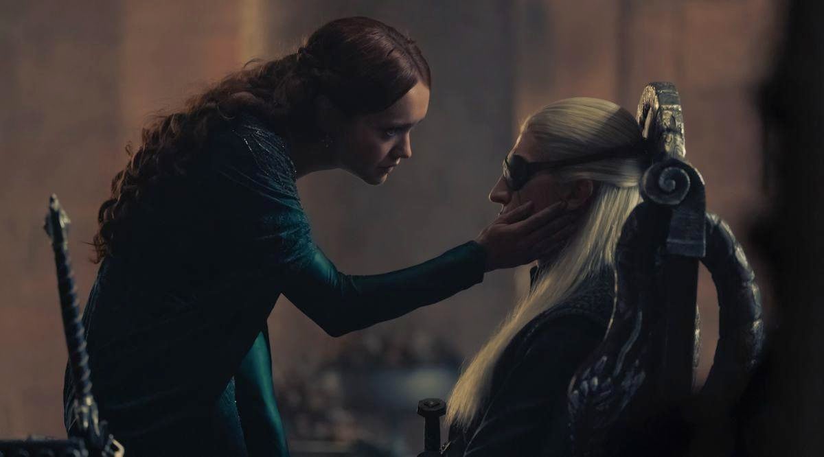 A woman with brown hair (Alicent) in a green dress leans over to touch the face of a man with long blonde hair and an eyepatch (Aemond).