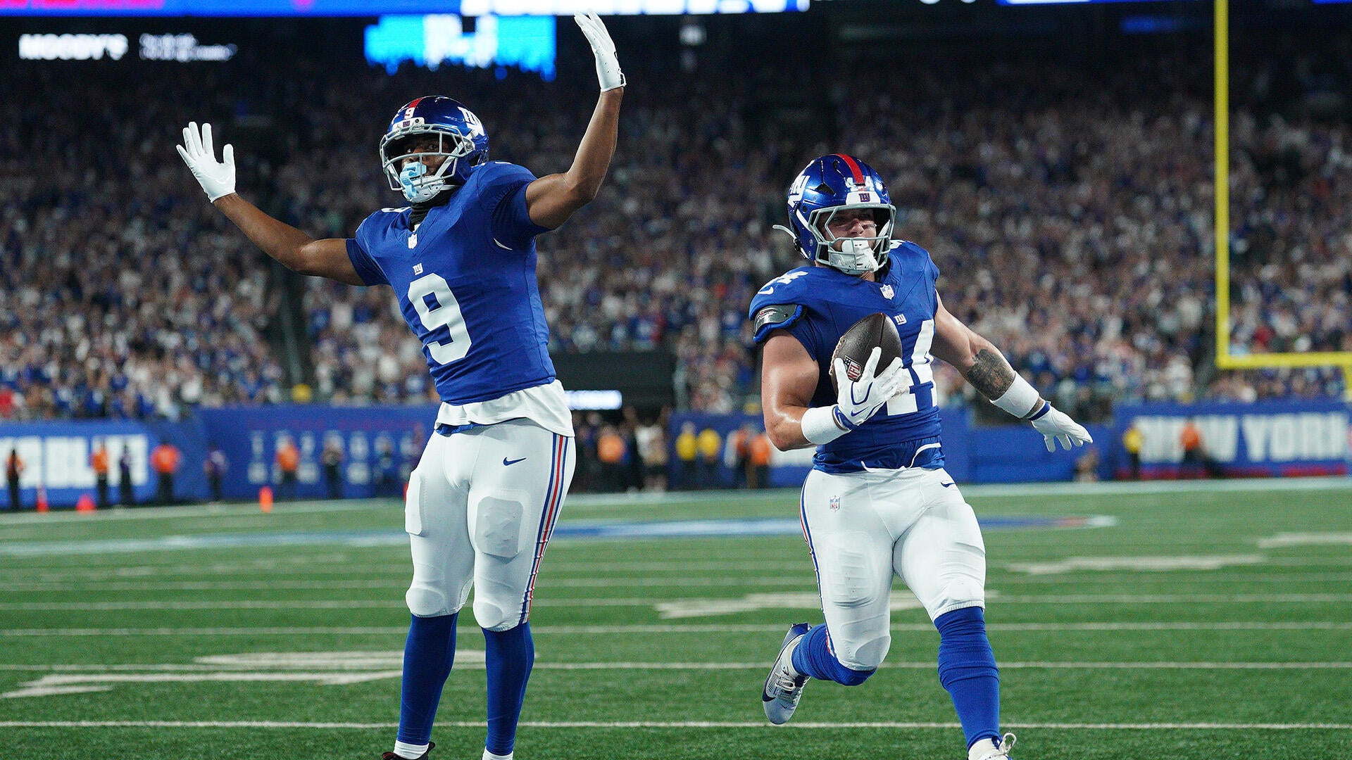 Two New York Giant players in blue jerseys celebrate as number 44 Cam Skattebo scores a touchdown.