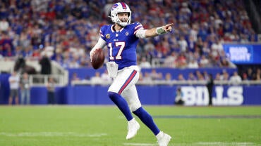 Bills QB Josh Allen, wearing a blue number 17 jersey, runs out of the pocket holding a football and points downfield.