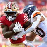 49ers RB, in a red jersey, carries the ball while a Broncos linebacker attempts to tackle him.