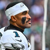 Eagles QB Jalen Hurts wears a white jersey and white sweatband on his head while looking out into the distance with his helmet off.