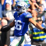 Seahawks running back wears a bright blue jersey, silver paints with a green and blue strip, and a silver helmet while scoring a touchdown.