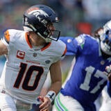 Broncos' Bo Nix, wearing a white number 10 jersey, runs with the football in his right hand while a Seahawks player chases him.