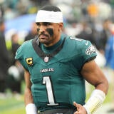 Eagles QB Jalen Hurts wears a white jersey and white sweatband on his head while looking out into the distance with his helmet off.