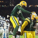 Josh Jacobs, in a green Packers jersey, celebrates his touchdown with his hands locked behind his back. Another Packers player jumps behind him to celebrate.