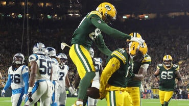 Josh Jacobs, in a green Packers jersey, celebrates his touchdown with his hands locked behind his back. Another Packers player jumps behind him to celebrate.